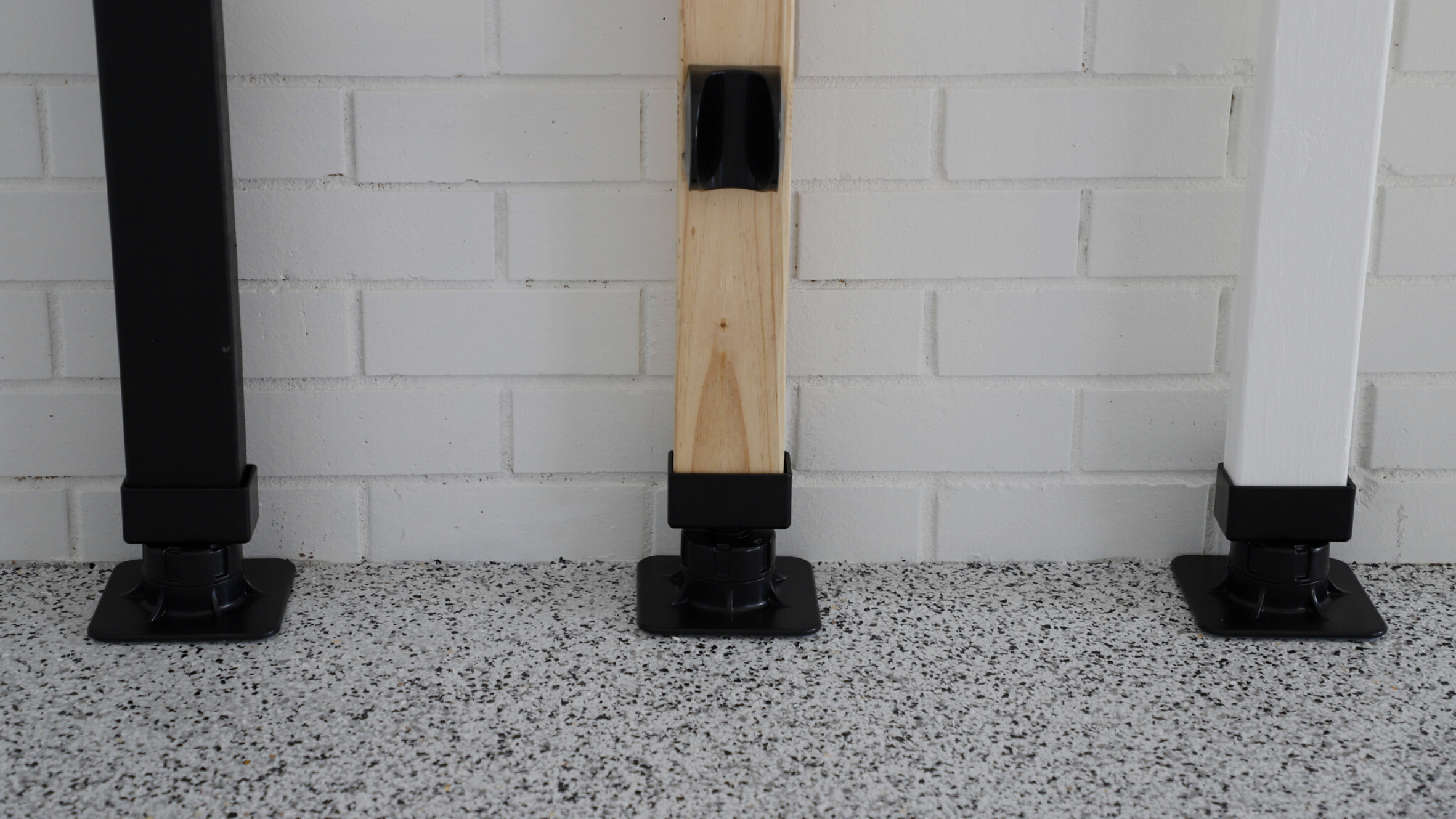 Three wooden posts with black brackets on a speckled floor against a white brick wall.