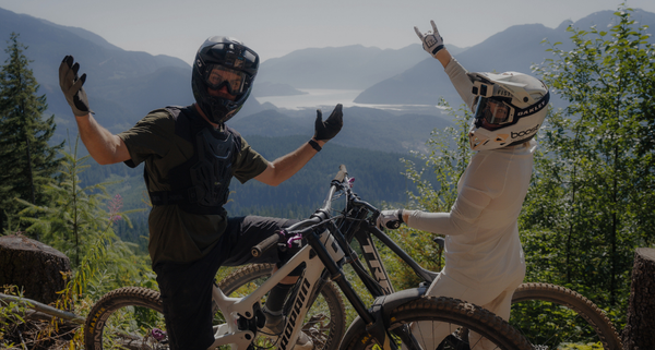 Two riders in helmets sitting on bikes on a cliff edge with mountains in the background